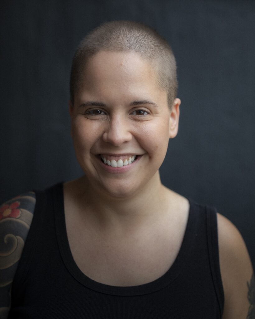 Foto da autora Bia Crespo. Ela é uma mulher branca, gorda, com a cabeça raspada e sorri abertamente para a foto. Usa uma regata preta e podemos ver uma tatuagem colorida no seu braço direito. O fundo é cinza. Os créditos da foto são de Renato Parada