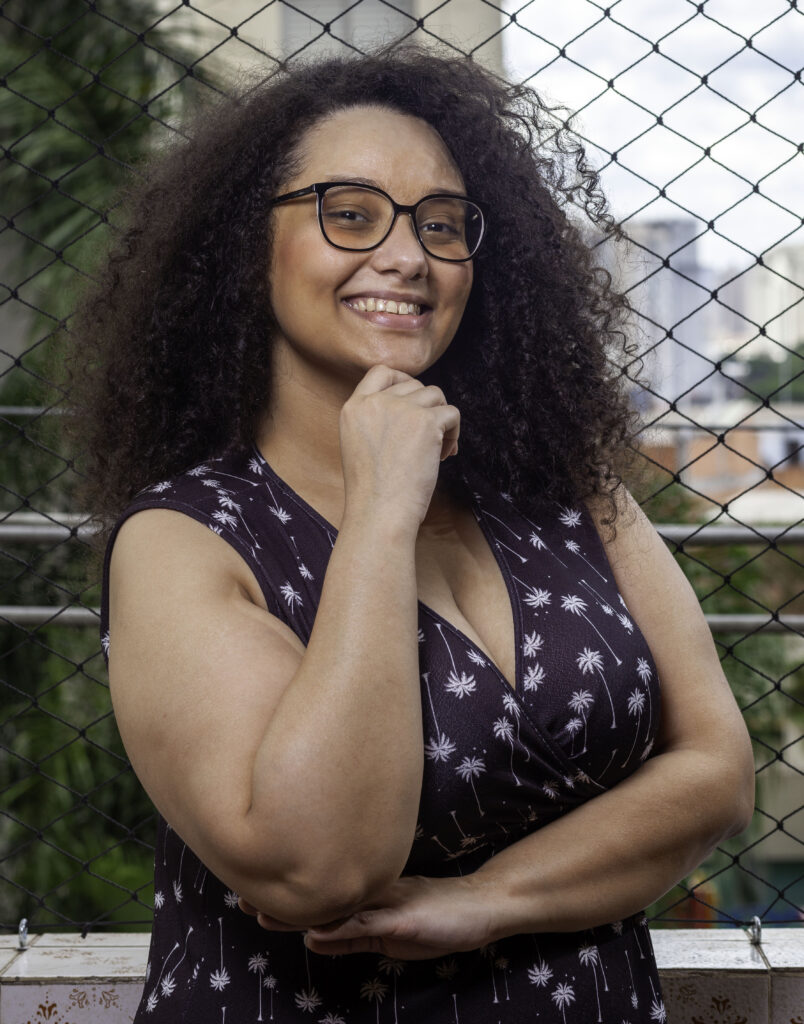 Foto da autora Solaine Chioro. Ela é uma mulher negra de pele clara, com cabelos crespos soltos. Usa óculos, está sorrindo para a foto, com a mão no queixo. Usa um vestido preto com flores brancas. Um dos braços está apoiando o outro, que tem a mão no queixo.